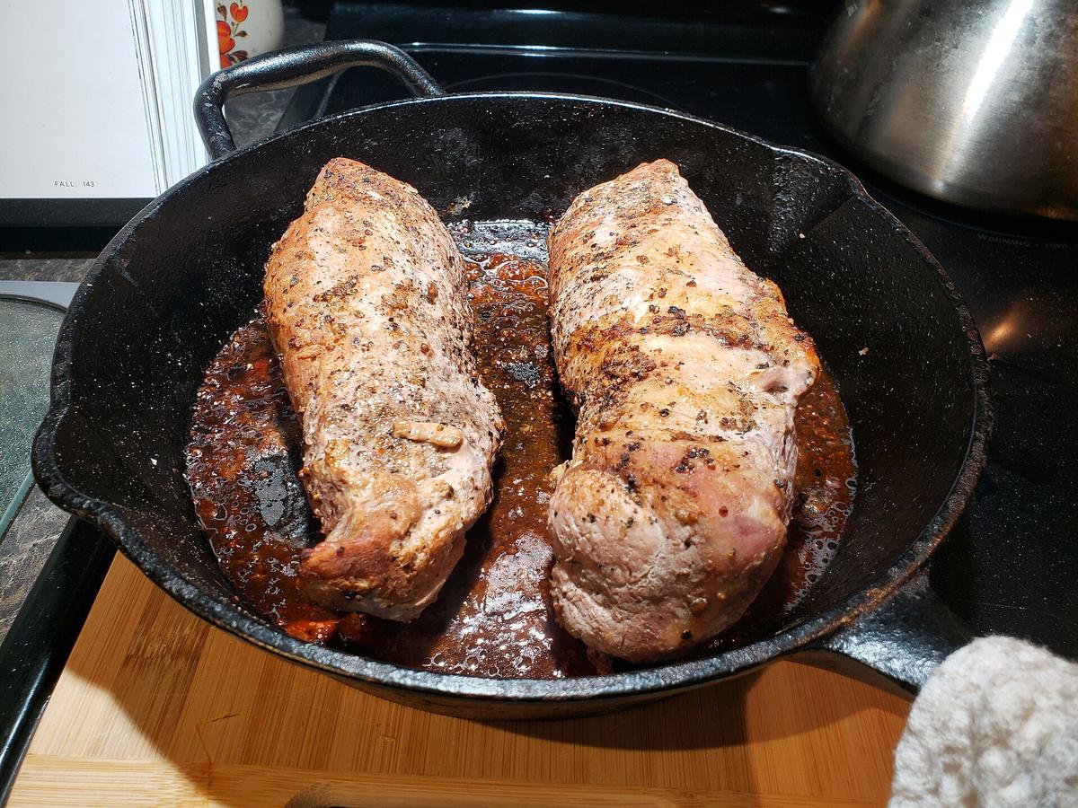 Two seasoned pork pieces searing in a cast iron pan on a stove