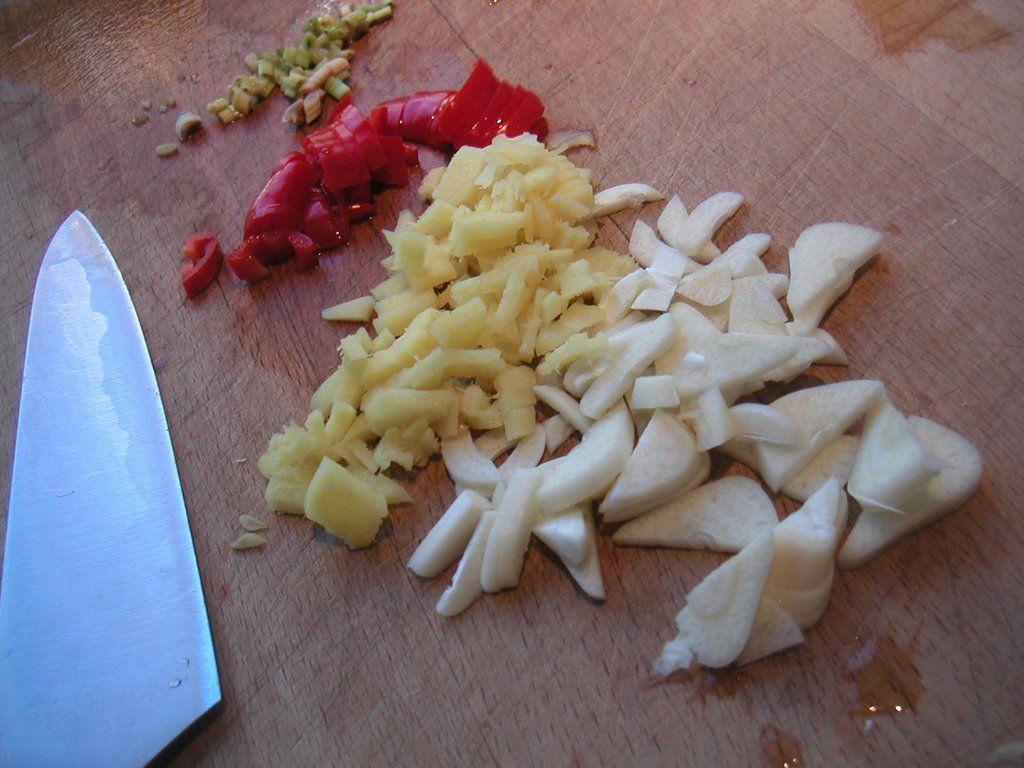 A chef's knife beside finely chopped garlic, ginger, and chili on a board